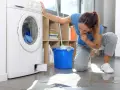 Donna preoccupata guarda la lavatrice che perde acqua e lascia residui di sporco sul pavimento.