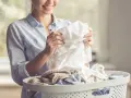 Donna sorride mentre controlla un capo bianco, temendo macchie grigie sui capi bianchi. Cesto di biancheria pieno.