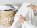 Mani che tengono un lenzuolo bianco, con macchie di candeggina visibili. Cesto di vimini sullo sfondo.