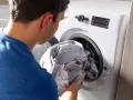 Uomo inserisce panni grigi nella lavatrice, selezionando il programma antimacchia per un bucato impeccabile.