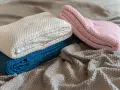 A pile of folded, knitted blankets in cream, blue, and pink, with a grey blanket spread out underneath.