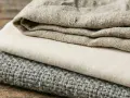 Tessuti impilati: lino grezzo, cotone chiaro e un tessuto grigio lavorato, che ricordano diversi tipi di velluto per la loro texture.