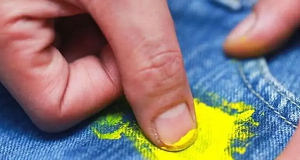 Dita che rimuovono macchia di vernice gialla dai jeans. Scopri come togliere tinta dai vestiti con metodi efficaci.