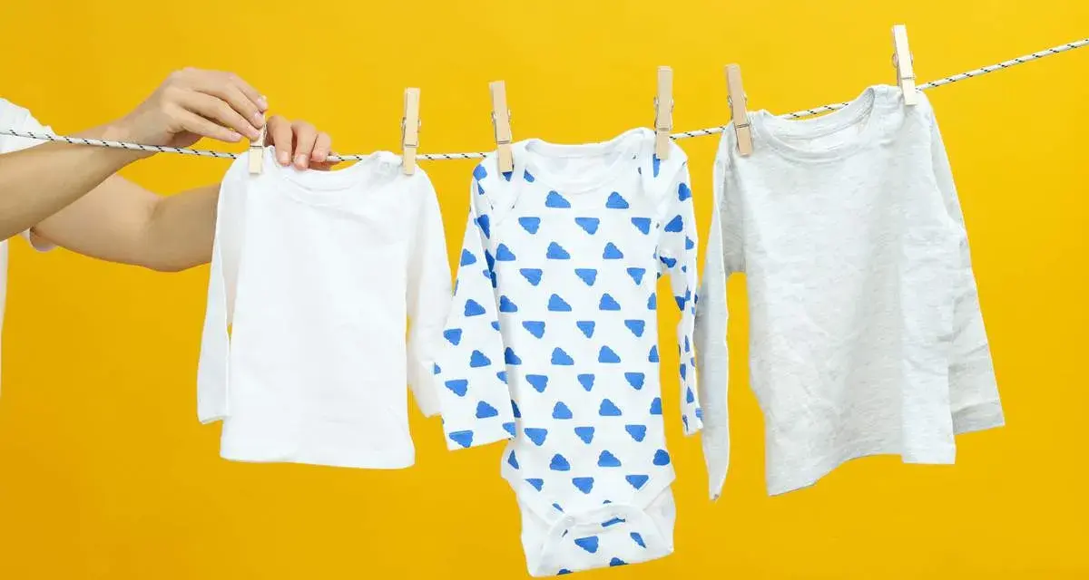 Mani che appendono tre body per neonato su uno stendibiancheria, uno bianco, uno con nuvole blu e uno grigio.