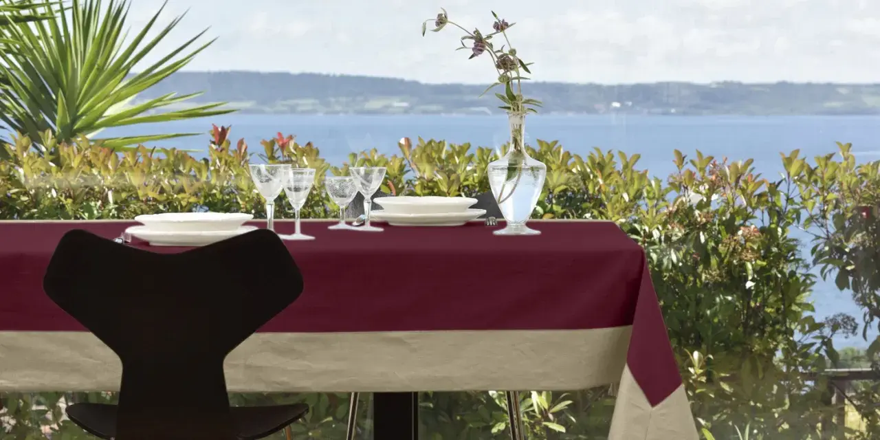 Tavola apparecchiata con tovaglia di lino bordeaux e beige, bicchieri, piatti e vaso con fiori. Un'idea su come rifinire una tovaglia di lino.