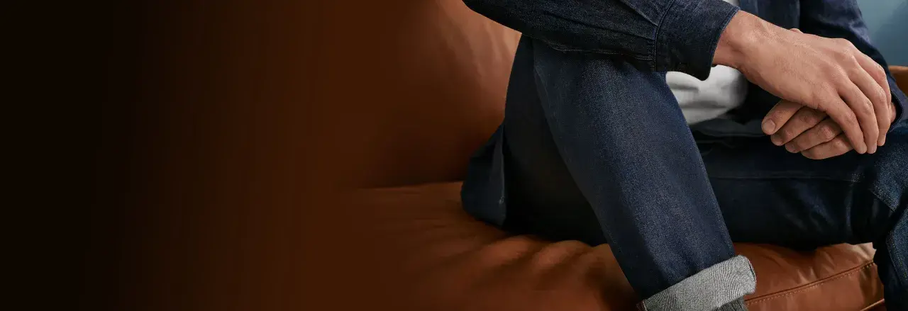 Uomo seduto su un divano di pelle, indossa un completo in denim. Il suo braccio è appoggiato sulla gamba, mostrando la texture del tessuto.