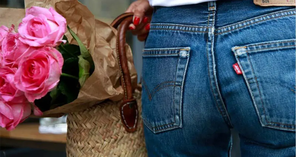 Un mazzo di rose rosa avvolto in carta marrone, appoggiato a una borsa di paglia. Indossa un paio di jeans dal colore jeans originale, con l'iconica etichetta rossa.
