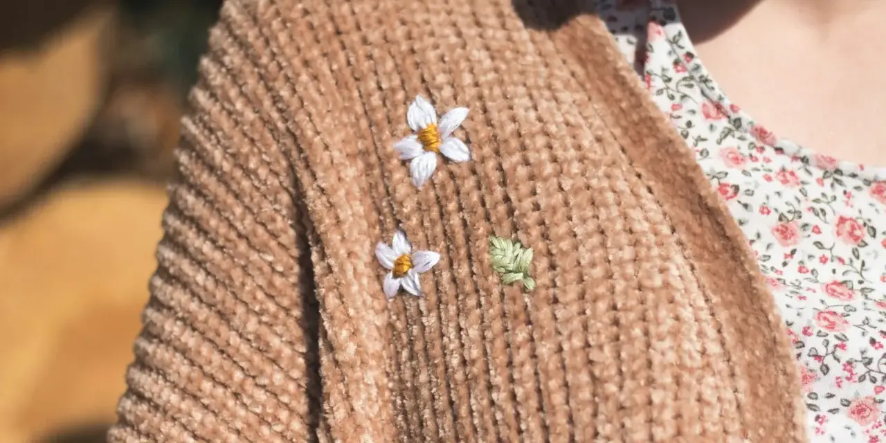 Delicati ricami su maglioni di lana: due fiorellini bianchi e una fogliolina verde aggiungono un tocco di natura a un caldo cardigan color caramello.