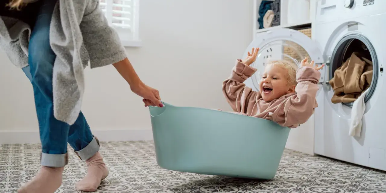 Bambina felice in una cesta, mentre una mano la aiuta a prepararsi per come lavare vestiti neonato. Lavatrice sullo sfondo.