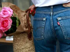 Un mazzo di rose rosa avvolto in carta marrone, appoggiato a una borsa di paglia. Indossa un paio di jeans dal colore jeans originale, con l'iconica etichetta rossa.