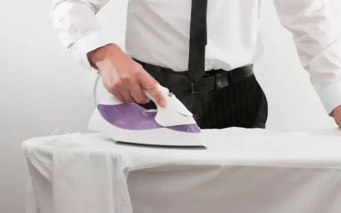 Uomo in camicia e cravatta stira una camicia bianca. L'immagine mostra l'uso di prodotti per stirare camicie, con il ferro da stiro in azione.