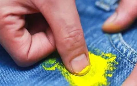 Dita che rimuovono macchia di vernice gialla dai jeans. Scopri come togliere tinta dai vestiti con metodi efficaci.