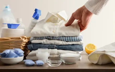 Mano indica una macchia su una camicia bianca, circondata da detersivi, bicarbonato per macchie di candeggina e limone.