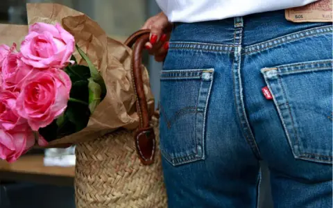 Un mazzo di rose rosa avvolto in carta marrone, appoggiato a una borsa di paglia. Indossa un paio di jeans dal colore jeans originale, con l'iconica etichetta rossa.