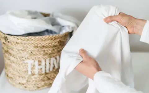 Mani che tengono un lenzuolo bianco, con macchie di candeggina visibili. Cesto di vimini sullo sfondo.