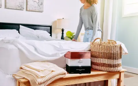 Donna sistema il letto con lenzuola bianche. Sul comodino, scatole di profumi e coperte. Scopri come ammorbidire il cotone nuovo.
