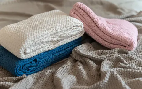 A pile of folded, knitted blankets in cream, blue, and pink, with a grey blanket spread out underneath.