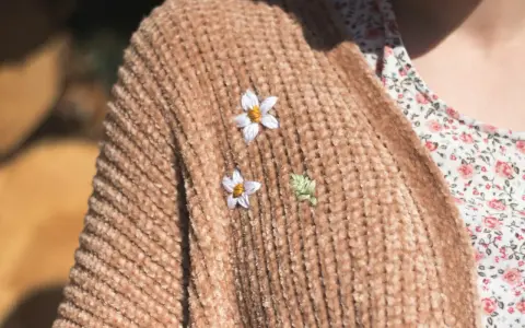 Delicati ricami su maglioni di lana: due fiorellini bianchi e una fogliolina verde aggiungono un tocco di natura a un caldo cardigan color caramello.