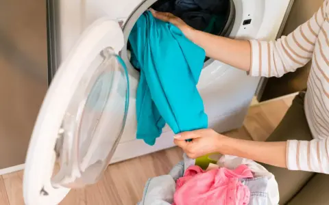 Mani che caricano una lavatrice con un capo di abbigliamento blu brillante. La cesta dei panni sporchi è piena di vestiti colorati, pronti per essere lavati.