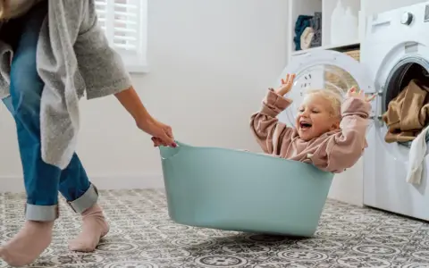 Bambina felice in una cesta, mentre una mano la aiuta a prepararsi per come lavare vestiti neonato. Lavatrice sullo sfondo.