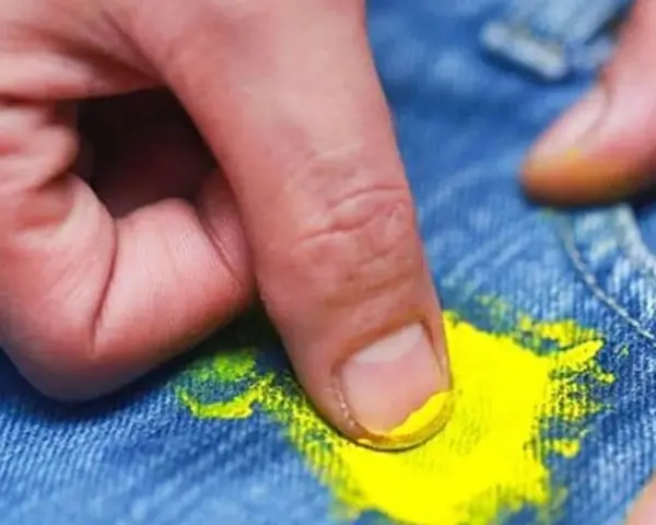 Dita che rimuovono macchia di vernice gialla dai jeans. Scopri come togliere tinta dai vestiti con metodi efficaci.