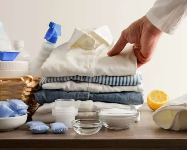 Mano indica una macchia su una camicia bianca, circondata da detersivi, bicarbonato per macchie di candeggina e limone.