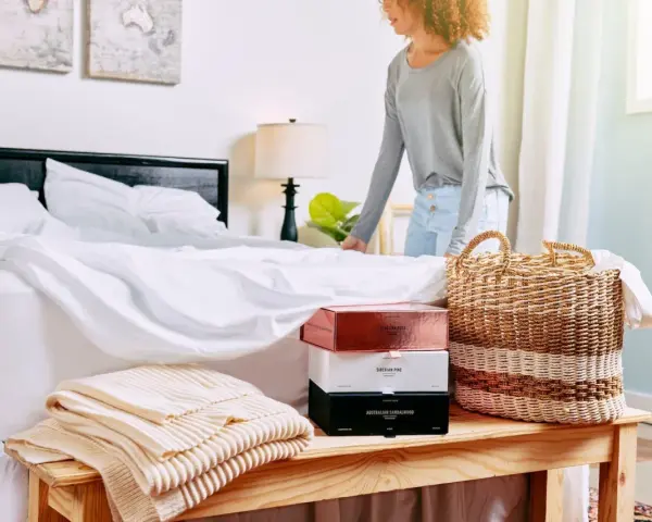 Donna sistema il letto con lenzuola bianche. Sul comodino, scatole di profumi e coperte. Scopri come ammorbidire il cotone nuovo.