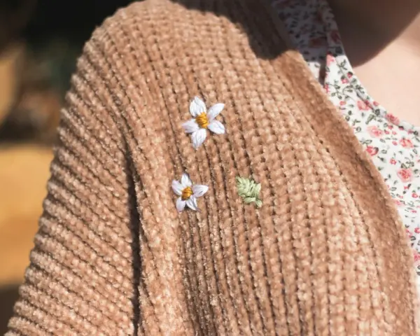 Delicati ricami su maglioni di lana: due fiorellini bianchi e una fogliolina verde aggiungono un tocco di natura a un caldo cardigan color caramello.