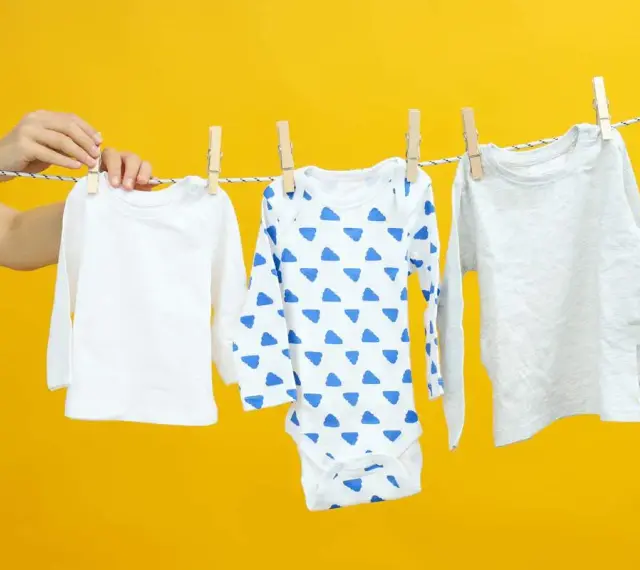 Mani che appendono tre body per neonato su uno stendibiancheria, uno bianco, uno con nuvole blu e uno grigio.