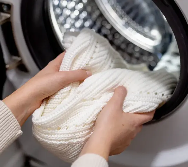 Mani che inseriscono un maglione bianco in lavatrice, per scoprire come sbiancare il bucato bianco in lavatrice e ottenere risultati perfetti.