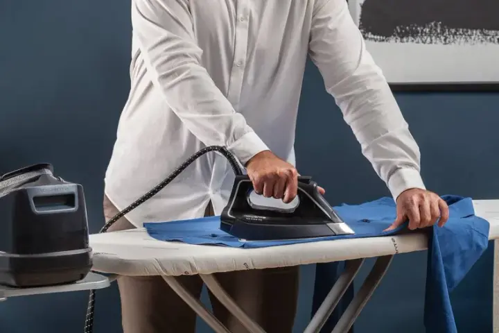Uomo stira una camicia blu su un asse da stiro. Impara come stirare il poliestere con cura per risultati impeccabili.