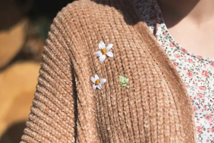 Delicati ricami su maglioni di lana: due fiorellini bianchi e una fogliolina verde aggiungono un tocco di natura a un caldo cardigan color caramello.
