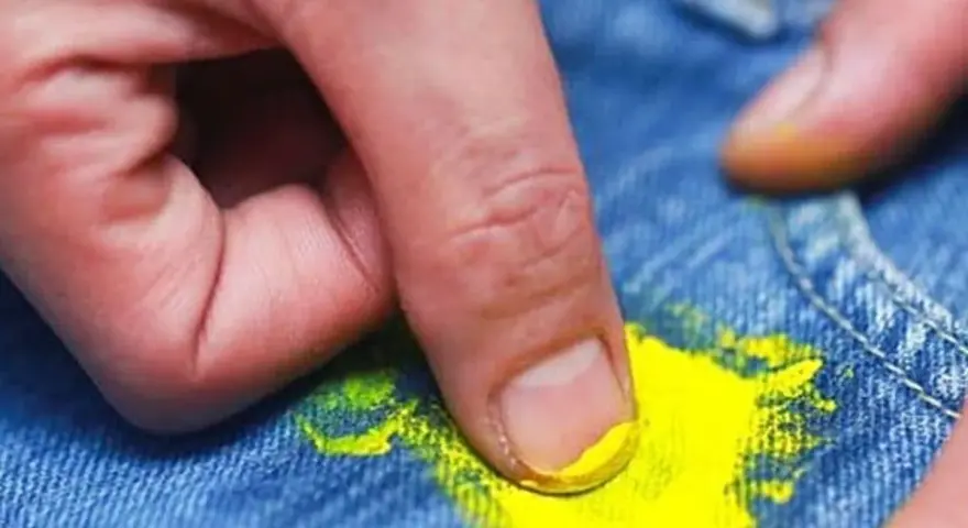 Dita che rimuovono macchia di vernice gialla dai jeans. Scopri come togliere tinta dai vestiti con metodi efficaci.