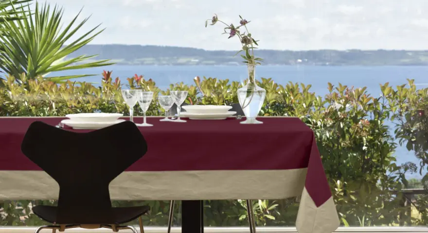 Tavola apparecchiata con tovaglia di lino bordeaux e beige, bicchieri, piatti e vaso con fiori. Un'idea su come rifinire una tovaglia di lino.