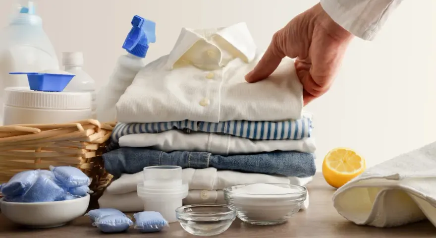 Mano indica una macchia su una camicia bianca, circondata da detersivi, bicarbonato per macchie di candeggina e limone.