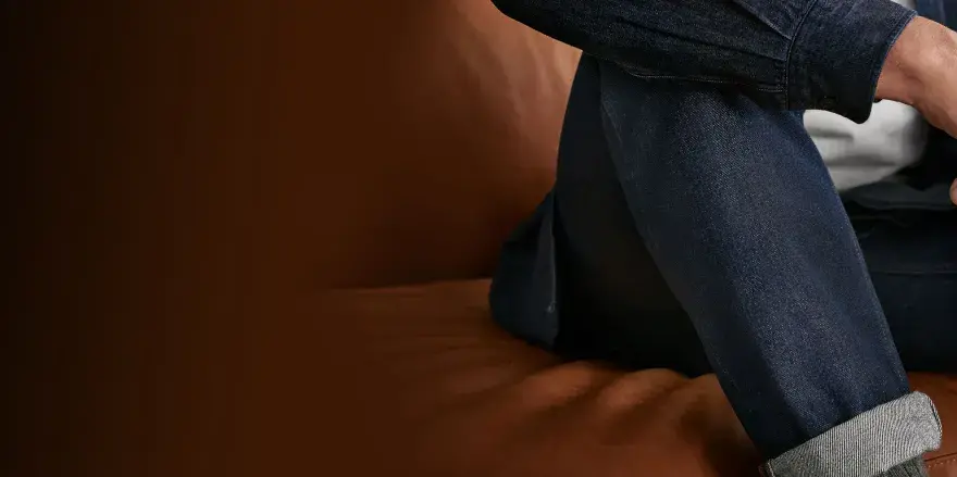 Uomo seduto su un divano di pelle, indossa un completo in denim. Il suo braccio è appoggiato sulla gamba, mostrando la texture del tessuto.