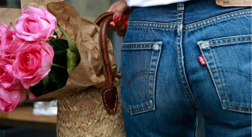 Un mazzo di rose rosa avvolto in carta marrone, appoggiato a una borsa di paglia. Indossa un paio di jeans dal colore jeans originale, con l'iconica etichetta rossa.