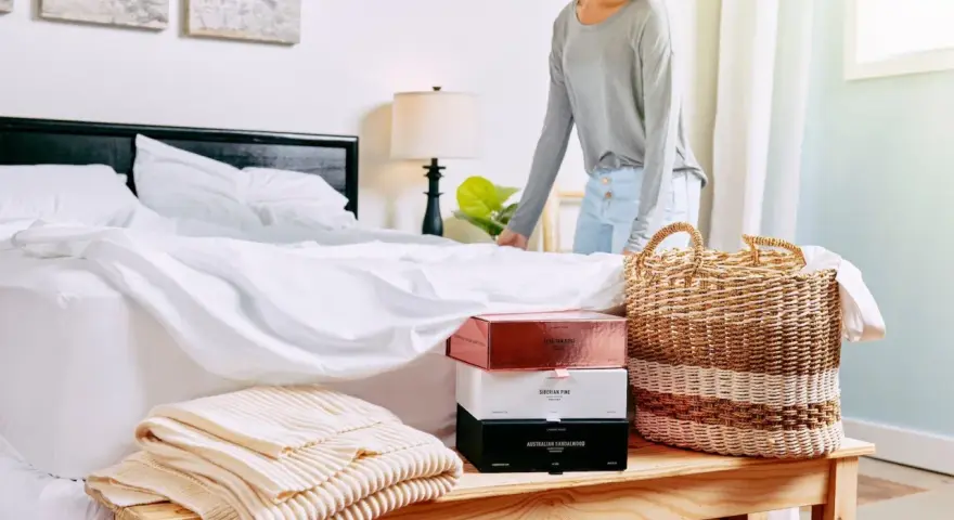 Donna sistema il letto con lenzuola bianche. Sul comodino, scatole di profumi e coperte. Scopri come ammorbidire il cotone nuovo.