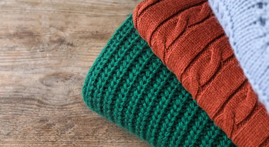 Close-up of folded knitted wool in green, rust, and light blue on a wooden surface.