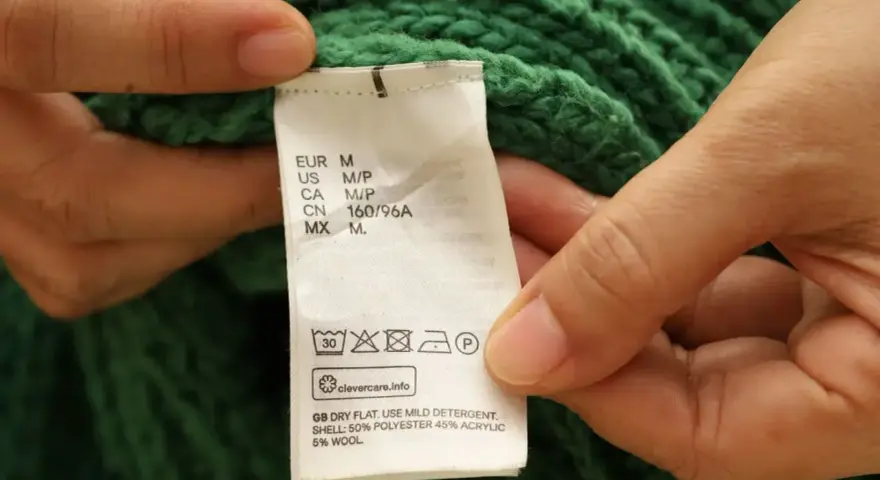 Etichetta di un maglione verde che indica il lavaggio in lavatrice a 30 gradi, con istruzioni per il lavaggio del cotone.