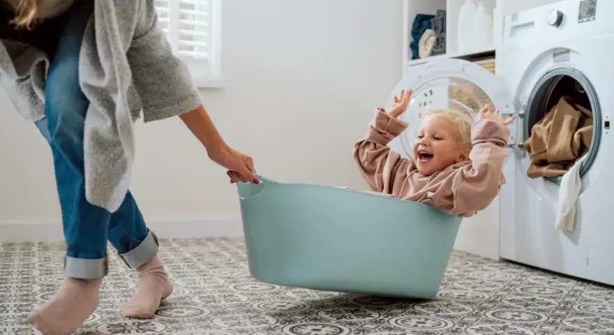 Bambina felice in una cesta, mentre una mano la aiuta a prepararsi per come lavare vestiti neonato. Lavatrice sullo sfondo.