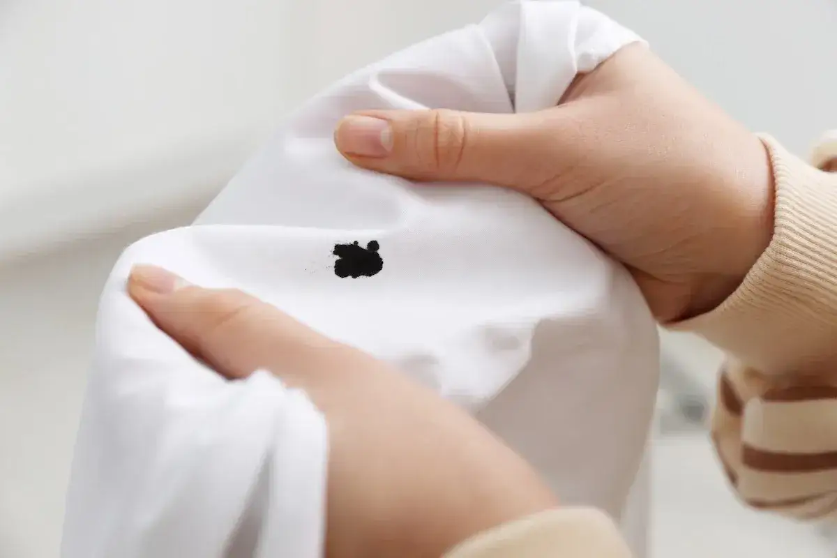 Mani che cercano di capire come togliere tinta dai vestiti, con una macchia nera su tessuto bianco.