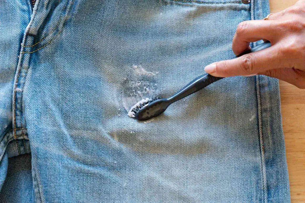 Una mano usa uno spazzolino per rimuovere una macchia di grasso dai jeans, mostrando un metodo efficace per come togliere il grasso dai jeans.