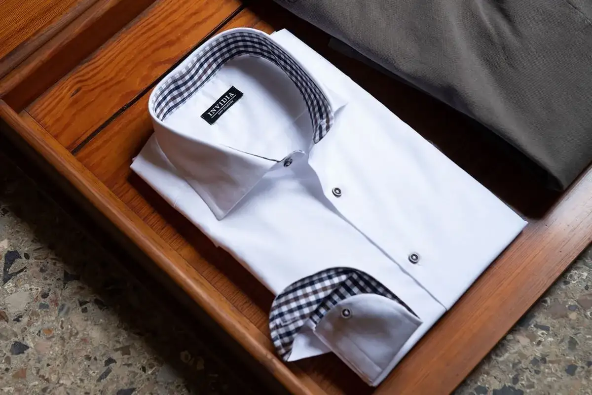 Camicia bianca piegata, pronta per essere stirata. Dettagli a quadretti sul colletto e polsino. Prodotti per stirare camicie perfetti per un look impeccabile.