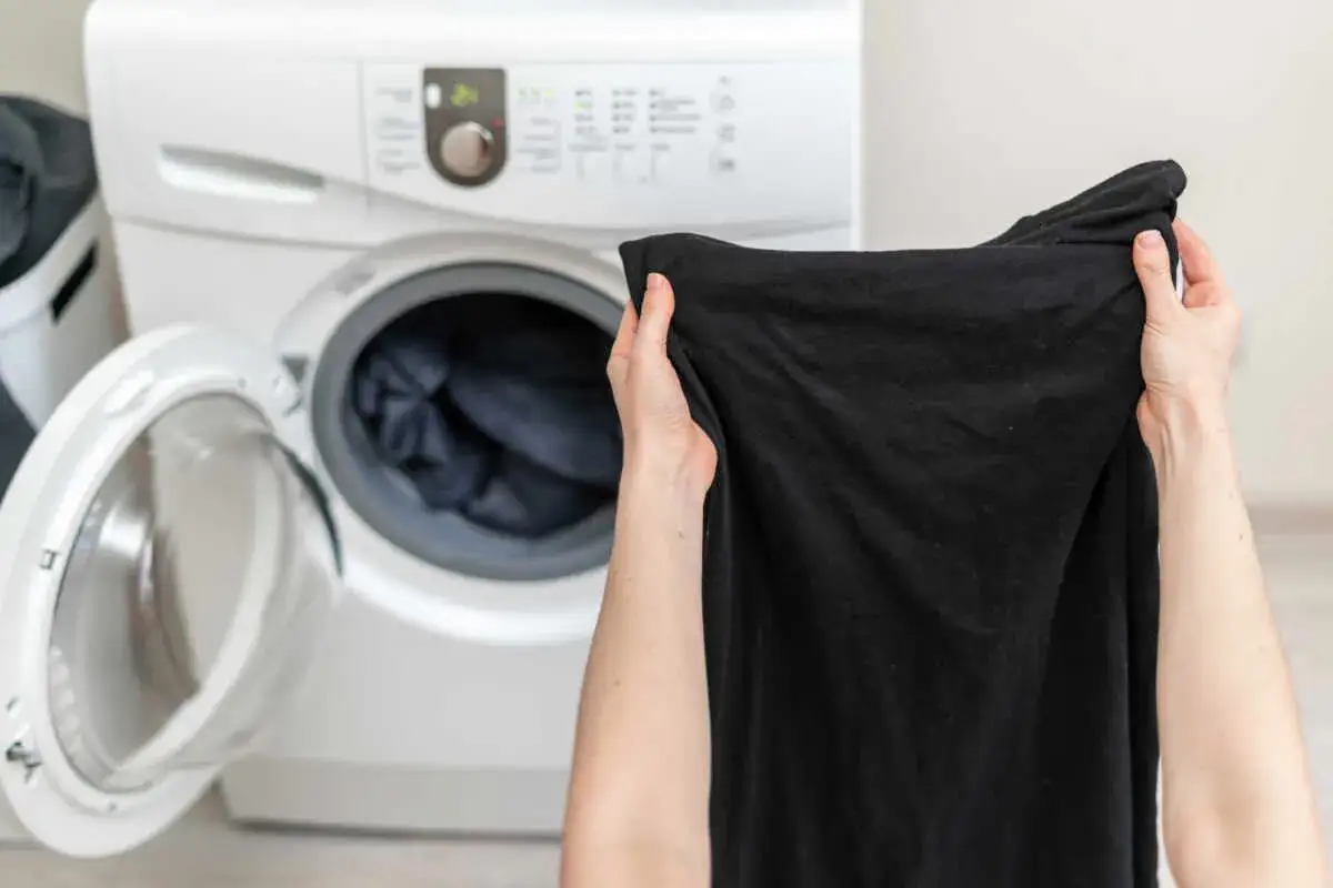 Mani che tengono un capo nero sbiadito, vicino a una lavatrice aperta. Rimedi della nonna per capi stinti.
