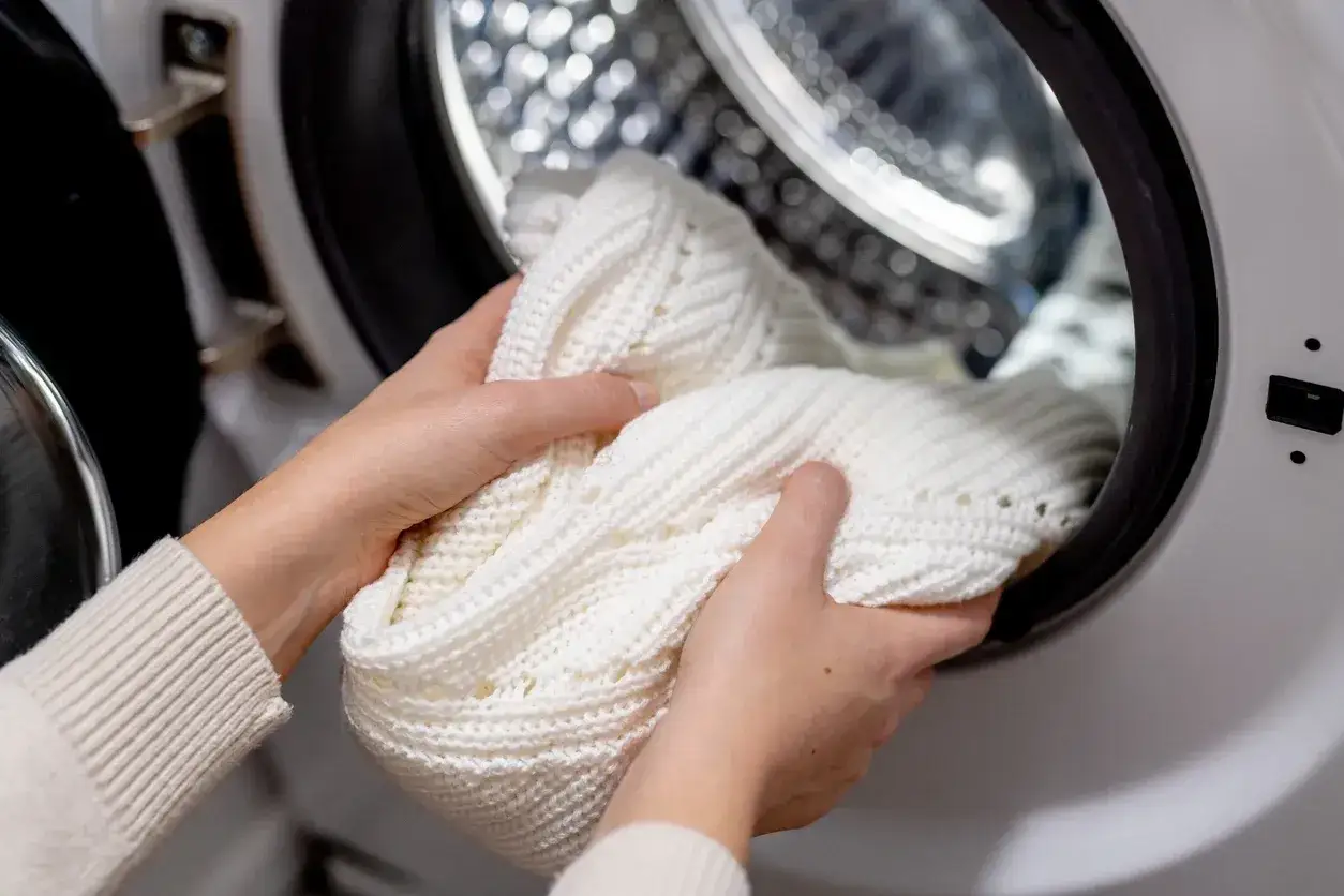 Mani che inseriscono un maglione bianco nella lavatrice. Speriamo che la lavatrice non lava bene i bianchi, altrimenti questo capo non tornerà candido.