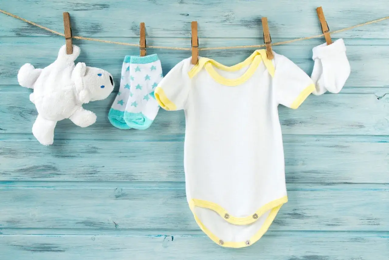 Come lavare vestiti neonato: body bianco, calzini azzurri a stelle, guantini e orsetto appesi su uno stendibiancheria.