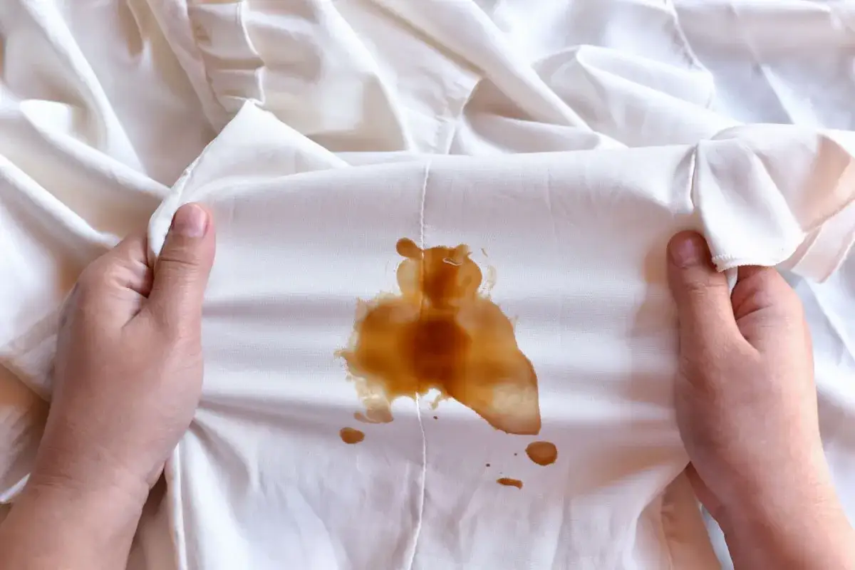 Mani che mostrano un vestito bianco macchiato di caff&egrave;, forse caduto da una borsa in pelle.