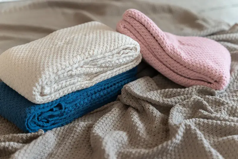 A pile of folded, knitted blankets in cream, blue, and pink, with a grey blanket spread out underneath.